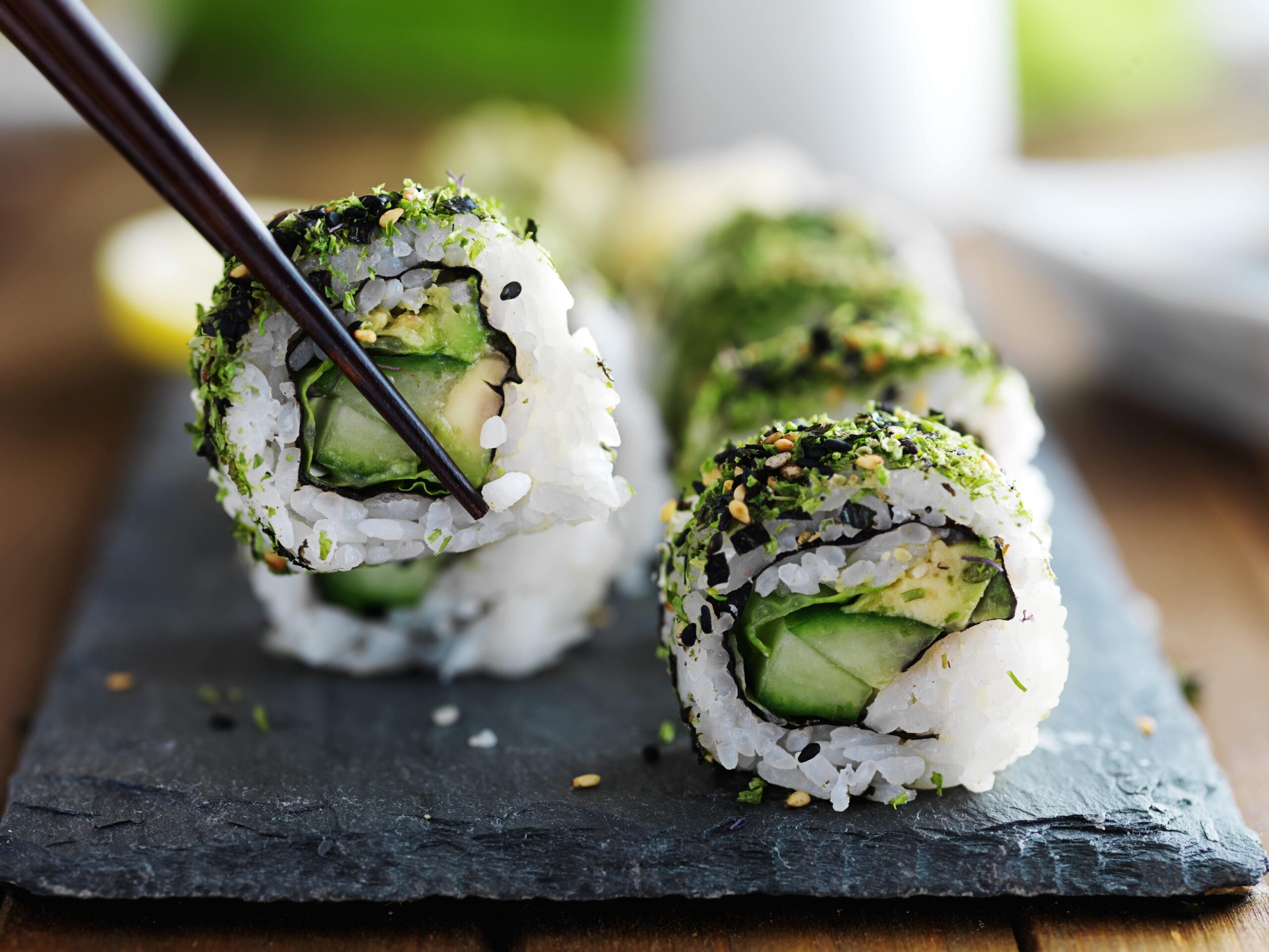 Nahaufnahme von veganen Sushi-Rollen mit Gurke und Avocado, bestreut mit Sesam und Kräutern. Eine Rolle wird mit Essstäbchen gehalten.