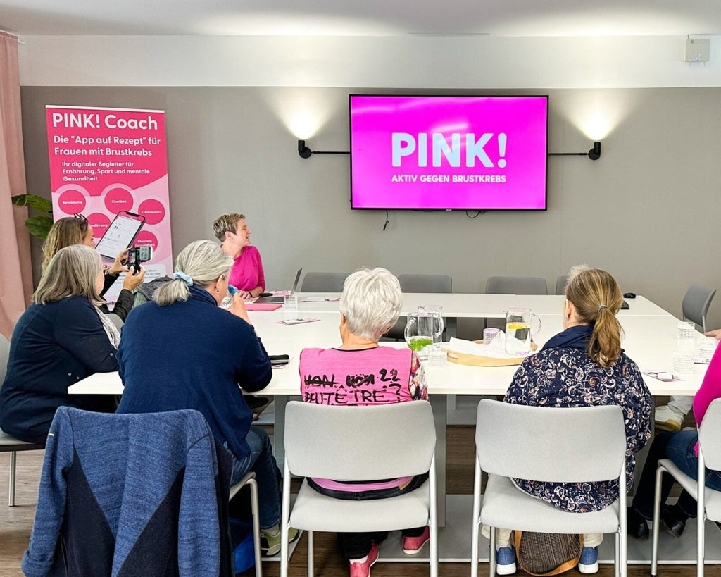 Mehrere Personen sitzen in einem Seminarraum an einem Tisch und hören einer Vortragenden zu. Auf dem Bildschirm und einem Banner ist der Schriftzug ‚PINK! Aktiv gegen Brustkrebs‘ zu sehen. Einige Teilnehmende tragen pinkfarbene Kleidung.